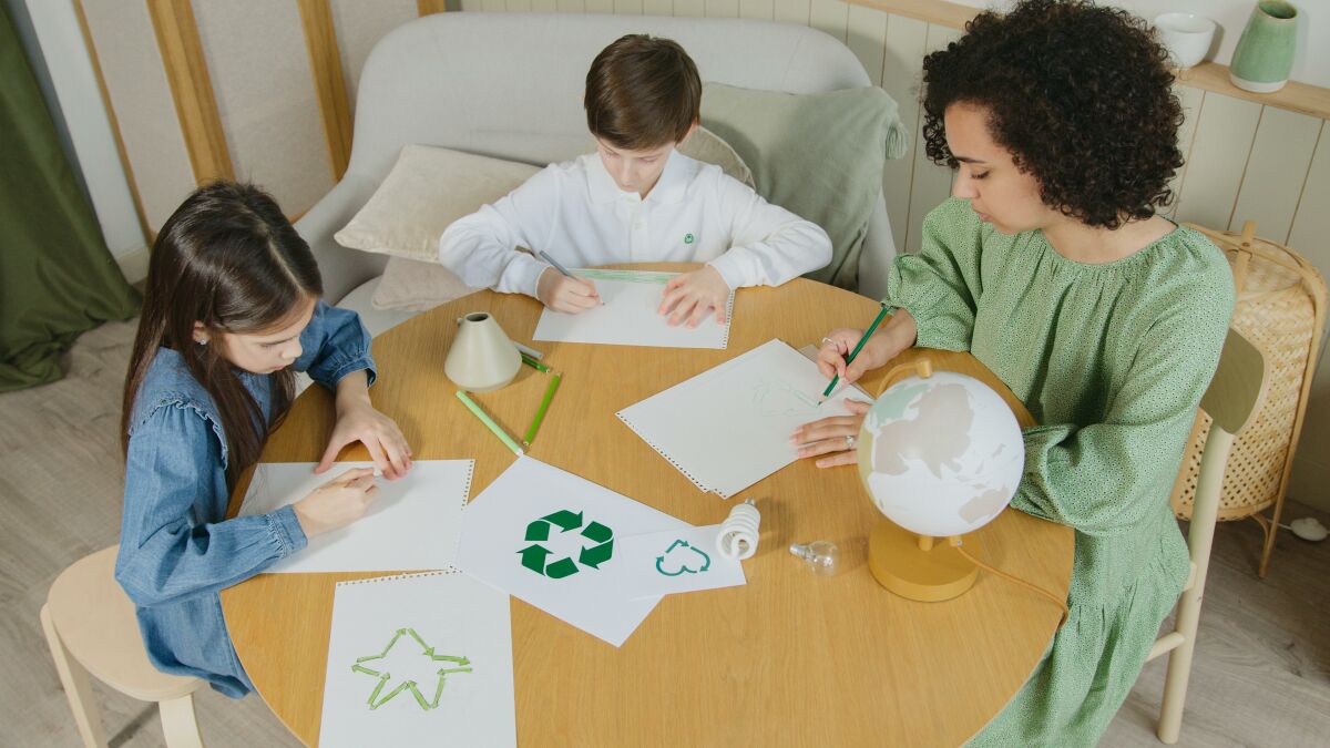 A woman and two children sitting at a round wooden table, drawing eco-friendly symbols such as recycling and stars with green pencils, promoting environmental education.